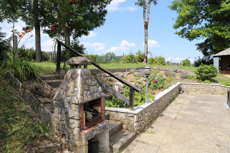 A stone outdoor fireplace with a wooden ramp leading up to it, surrounded by a stone wall, greenery, and a paved pathway.