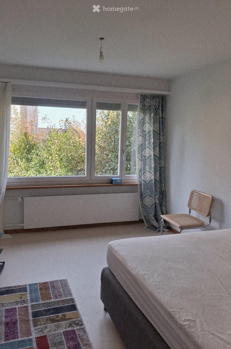 Bedroom with a bed, a chair, a radiator, and a window with curtains. The room has light-colored walls and carpeted flooring.