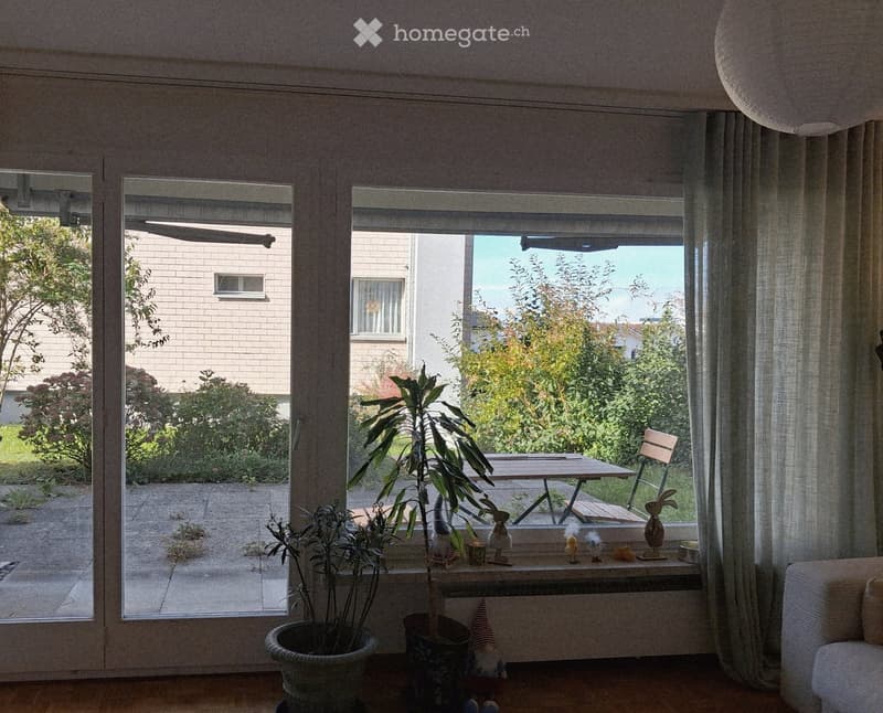 Bright living room with large sliding glass doors opening to a patio, featuring a sofa, a small table with chairs, and potted plants near the window.