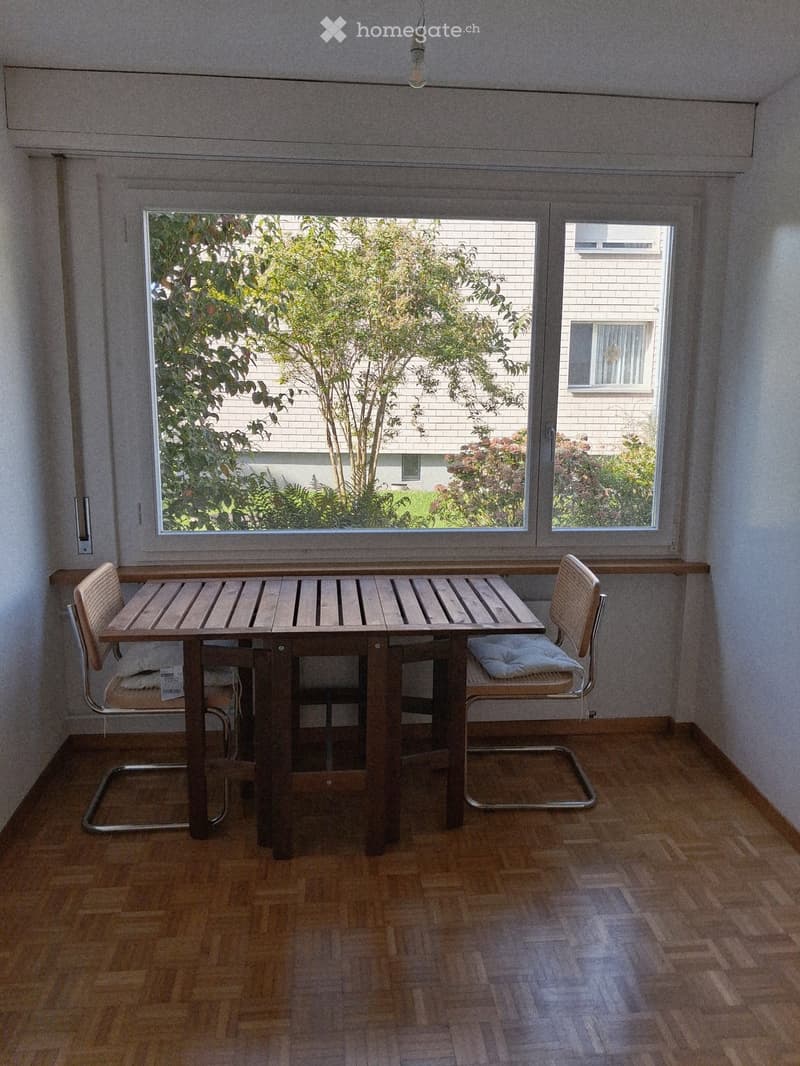 A small, furnished room with wooden flooring, a wooden table, and two chairs near a large window offering a view of greenery and neighboring buildings.