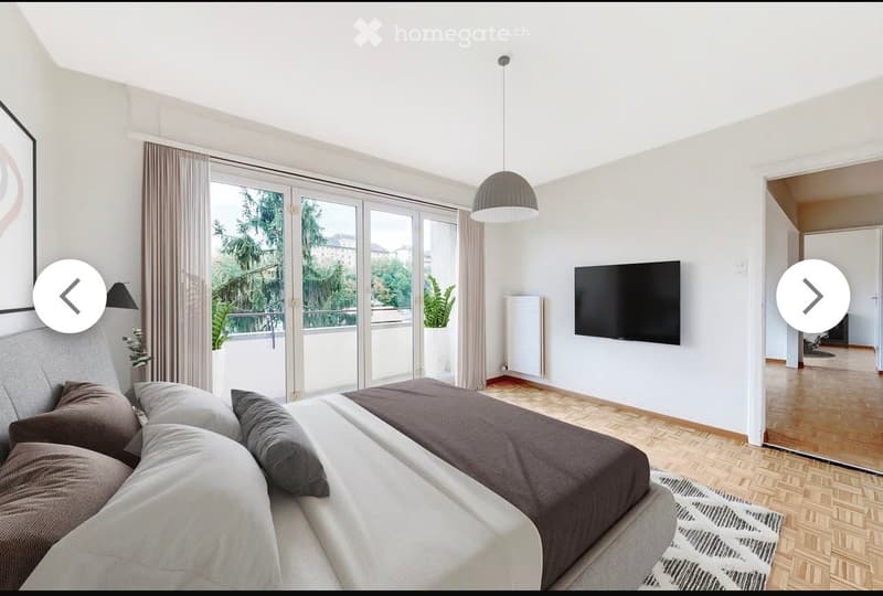 Modern bedroom with a large bed, pillows, a television mounted on the wall, a pendant light, and a view of greenery through large windows.