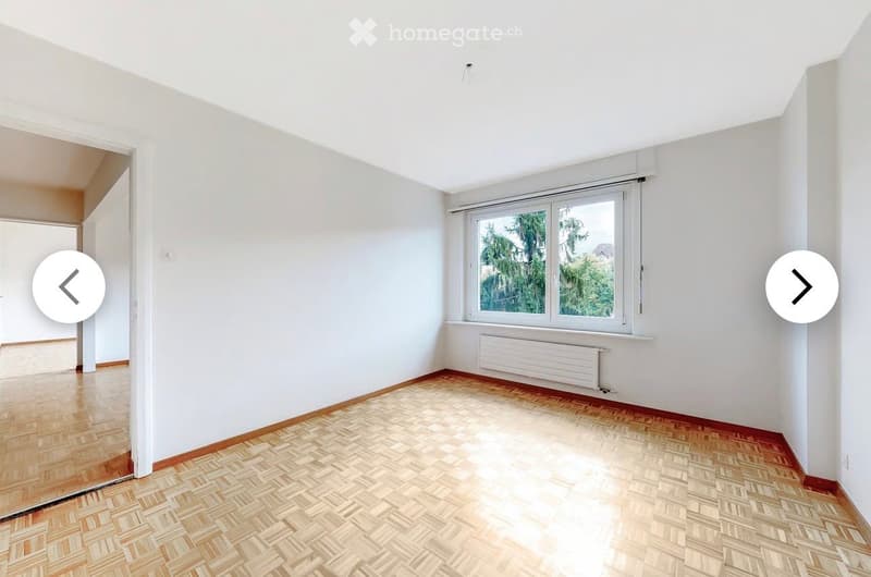 Empty room with wooden parquet flooring, white walls, large window with white frame, radiator below window, and a door leading to another space.