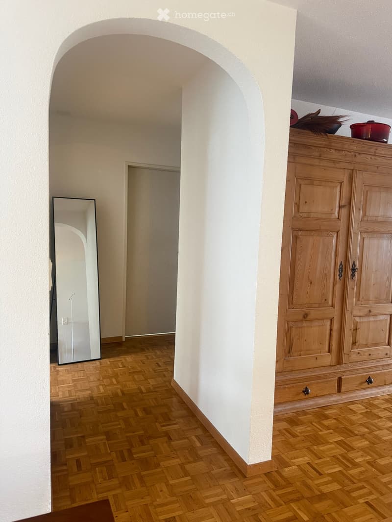 A hallway with wooden herringbone flooring, white walls, and an arched doorway leading to another room. A large mirror is mounted on the left wall, and a wooden cabinet with drawers and doors is on the right side.