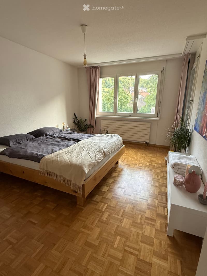 Cozy bedroom with wooden herringbone flooring, large window with view, bed with dark bedding, nightstand with lamp, potted plants, radiator, and a small table with decorative items.