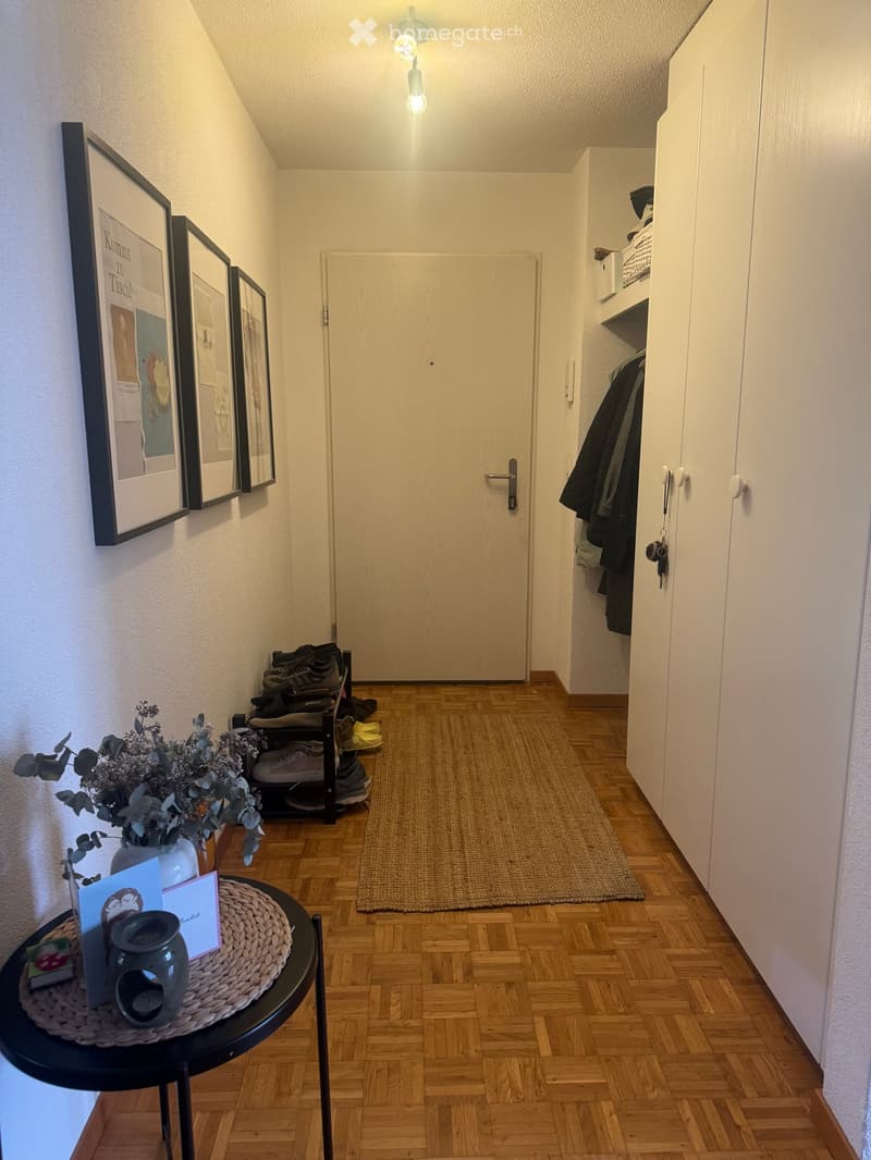 Narrow hallway with wooden flooring, a small round table with a candle and cards, a shoe rack with shoes, a coat closet, and framed artwork on the wall.