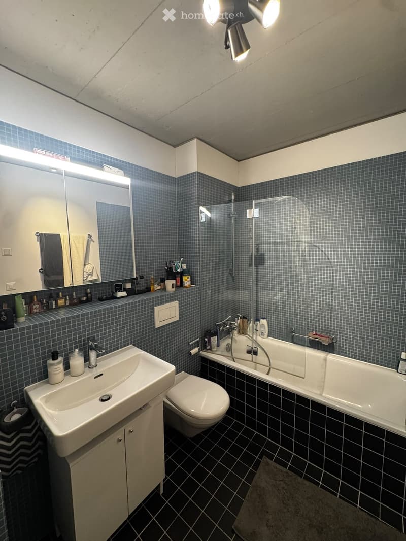 Modern bathroom with blue tiled walls, black tiled floor, white sink, toilet, bathtub, and mirror. Shelves hold toiletries and towels.