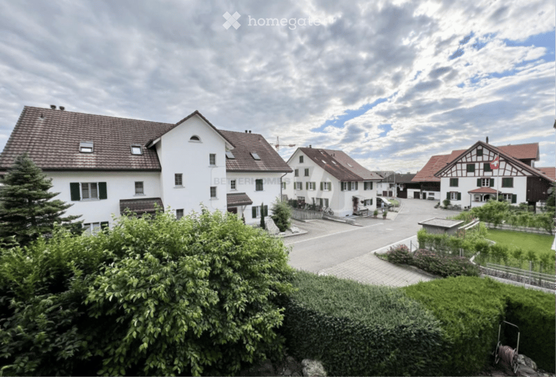 A row of traditional Swiss houses with pitched roofs and white facades, surrounded by greenery and a paved road.