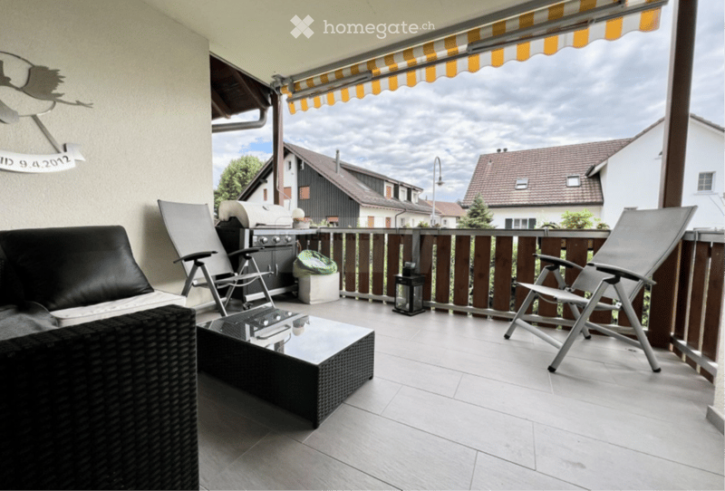 Balcony with a black leather sofa, wicker coffee table, two folding chairs, and a barbecue. The balcony has a wooden railing and a striped awning.
