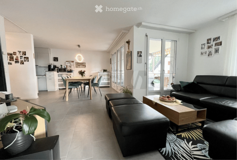 Open-plan living and dining area with a black leather sofa, wooden coffee table, dining table with chairs, and a small kitchen area with cabinets and appliances. Large sliding glass doors lead to a balcony.