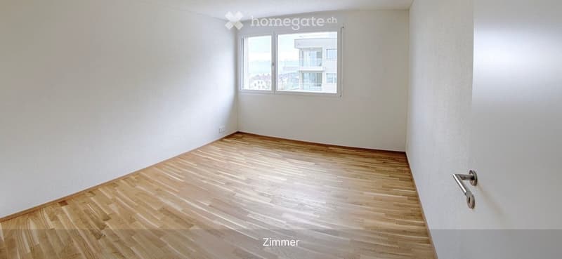 Empty room with light wooden flooring and white walls, featuring a large window with a view of a building and outdoor area.