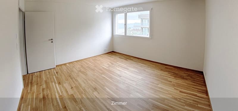 Empty room with light wooden flooring, white walls, and a large window offering a view of the sea and buildings.