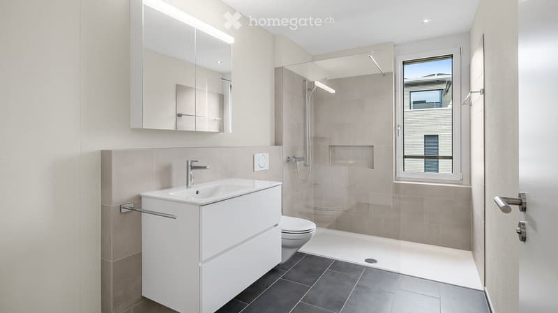 Modern bathroom with a white sink, mirror, toilet, and a glass-enclosed shower area. The room features beige tiles and a large window offering a view.