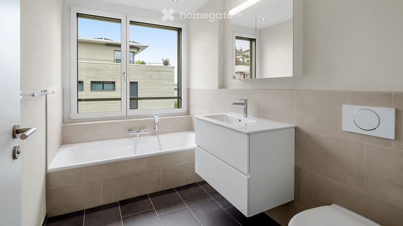Modern bathroom with a bathtub, sink, mirror, and toilet. Features large windows with a view of the building exterior.