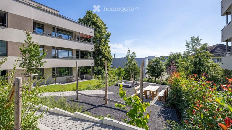 Modern residential complex with a landscaped garden area, featuring a patio with a wooden table and benches, surrounded by greenery and trees. The building has large windows and balconies.