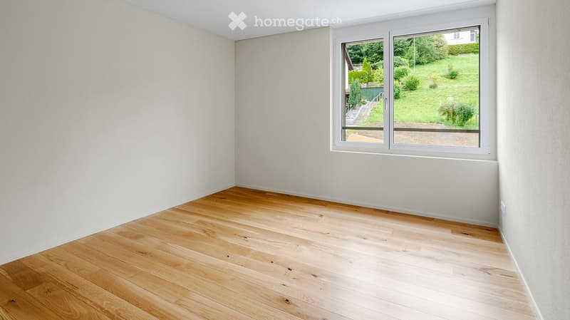 Empty room with light wooden flooring and white walls, featuring a large window with a view of a green garden.