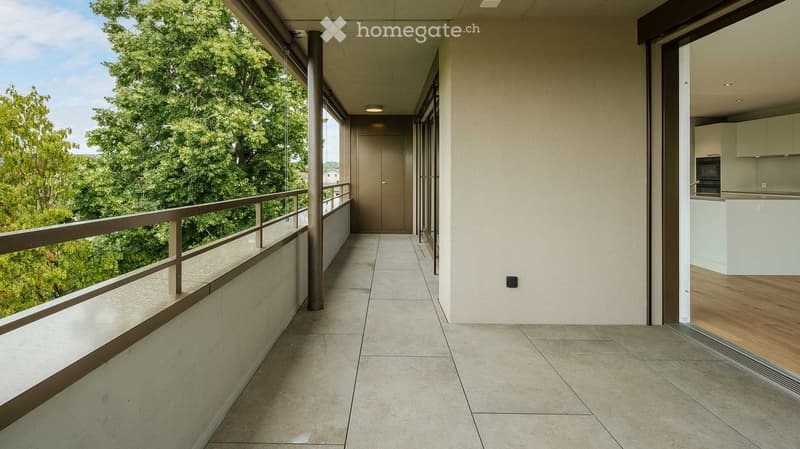 Modern balcony with tiled flooring, metal railing, and a view of lush greenery. An open doorway leads into a contemporary kitchen area with white cabinetry and wooden flooring.
