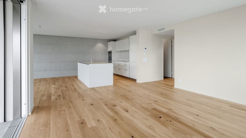 Open-plan kitchen and living area with light wooden flooring, white cabinetry, and a central island. The space is unfurnished, with no visible furniture or appliances.