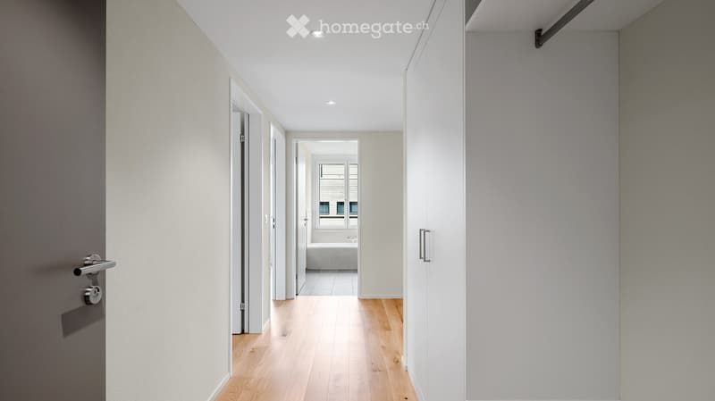 Modern hallway with light wooden flooring, white walls, recessed ceiling lights, and open doors leading to other rooms.