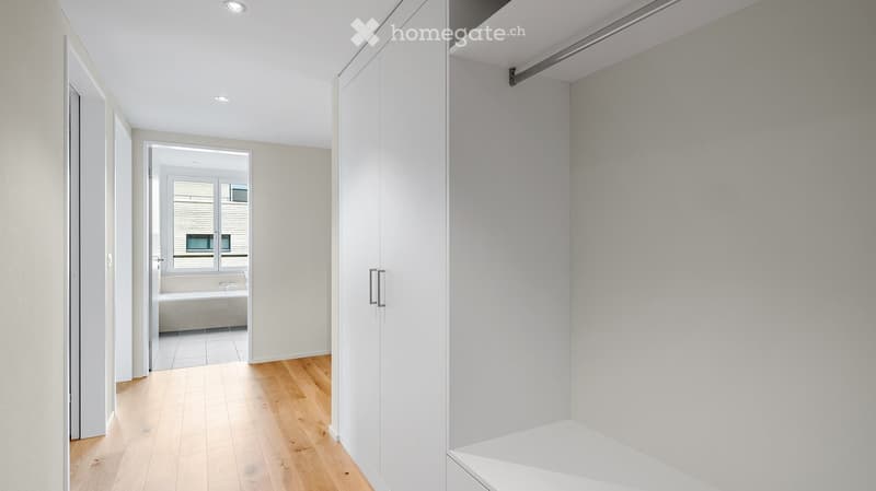 Empty hallway with light wooden flooring, white walls, and built-in closet with sliding doors. The hallway leads to a room with a bathtub visible through an open doorway.