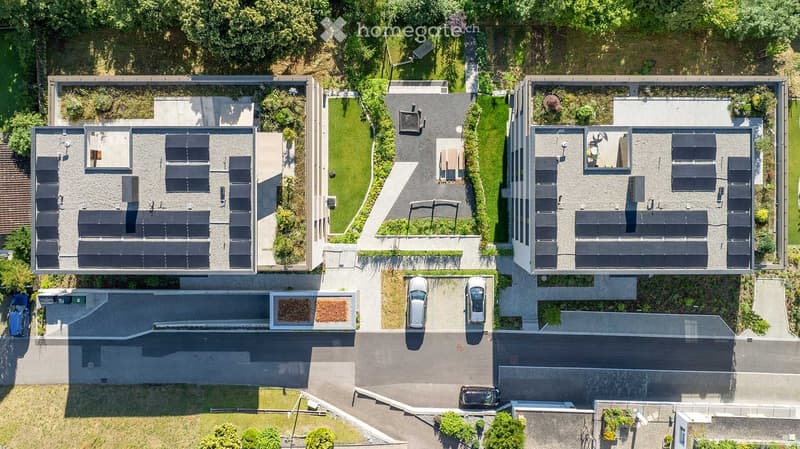 Aerial view of a modern residential building with multiple solar panels on the roof, surrounded by greenery and parking spaces.