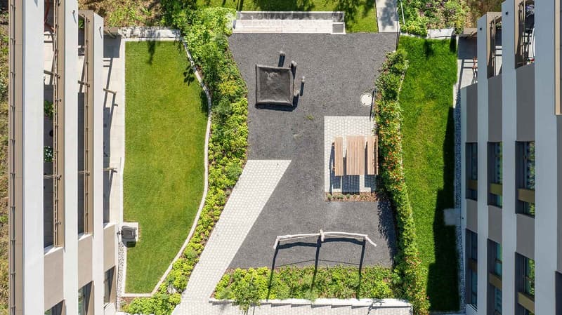 A well-maintained garden area with a central lawn, surrounded by paved pathways and wooden benches, located between two modern buildings.