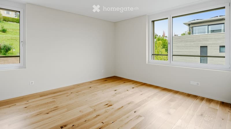 Empty room with light wooden flooring and white walls, featuring large windows with views of greenery and neighboring buildings.