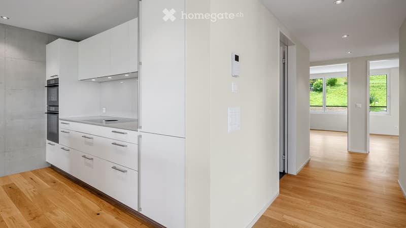 Modern kitchen with white cabinets, stainless steel appliances, and wooden flooring. The kitchen is connected to a bright living area with large windows and wooden flooring.