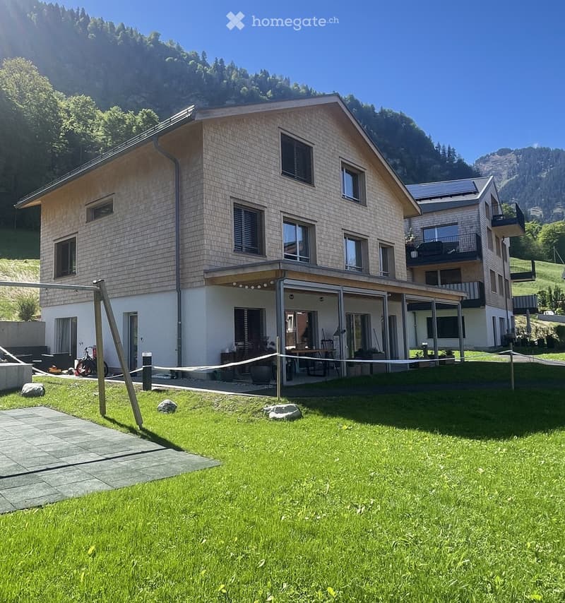Two-story modern house with light-colored siding, large windows, and a spacious covered patio area. The house is surrounded by a well-maintained lawn and is situated in a scenic, mountainous area.
