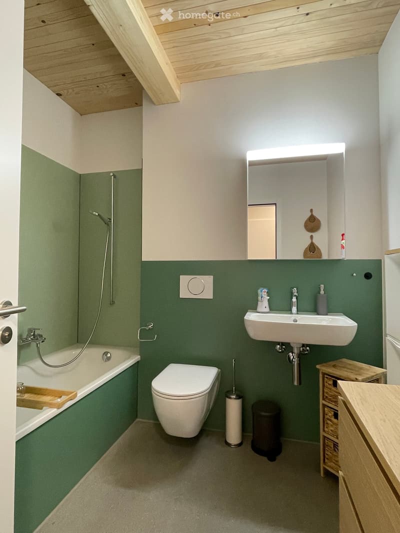 Modern bathroom with green lower walls, white upper walls, and a sloped wooden ceiling. Features a bathtub, toilet, sink with a mirror, and a small wooden stool with a woven basket.