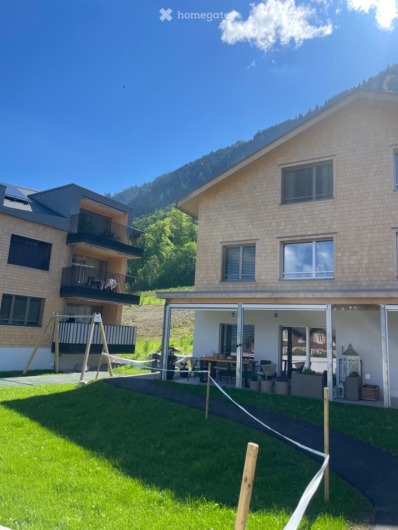 A modern residential complex with a grassy lawn, patio furniture, and a swing set, surrounded by trees and mountains.