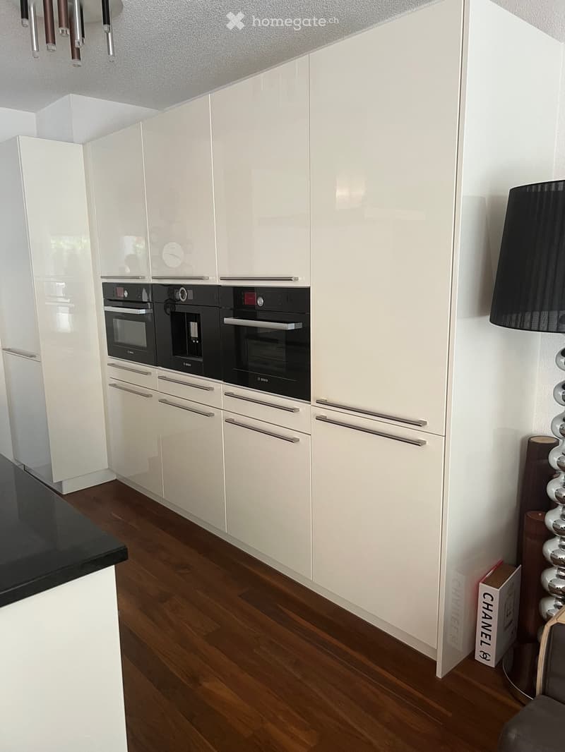 Modern kitchen with white cabinets, stainless steel handles, and built-in ovens. Wooden flooring and a black countertop are visible.