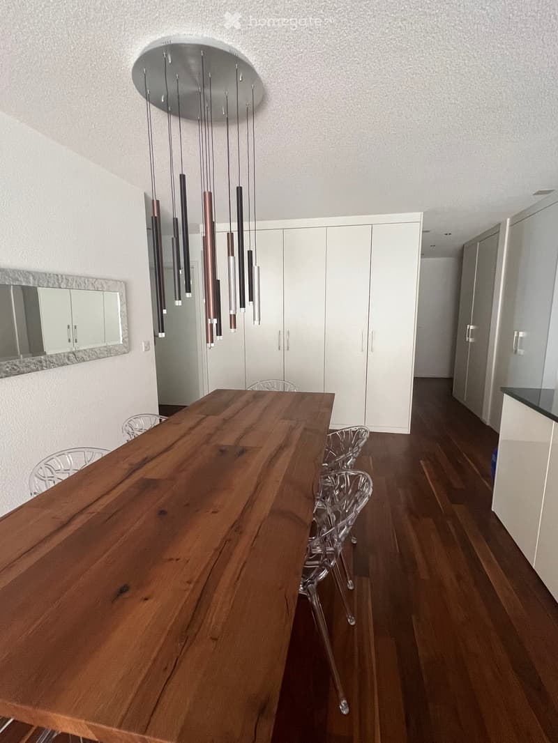 Modern dining area with a large wooden table, transparent chairs, white cabinets, and a decorative hanging light fixture.