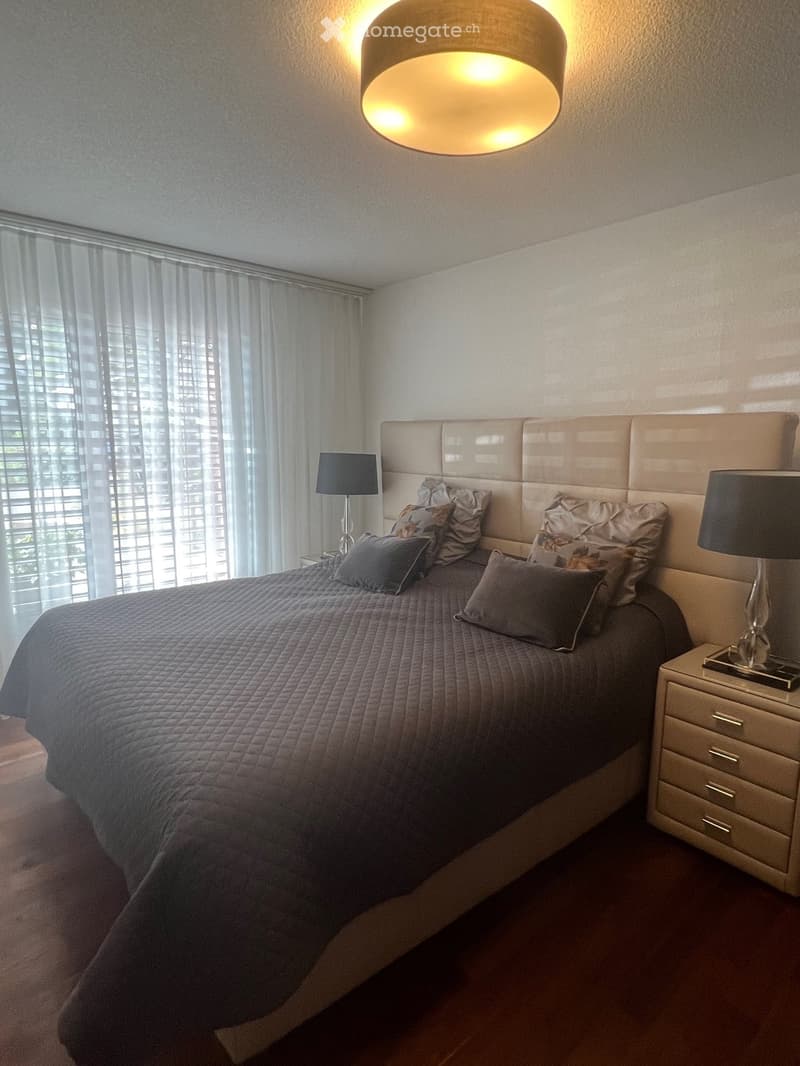 Modern bedroom with a large bed, headboard, nightstand, and lamps. The room features large windows with blinds and a warm, ambient ceiling light.