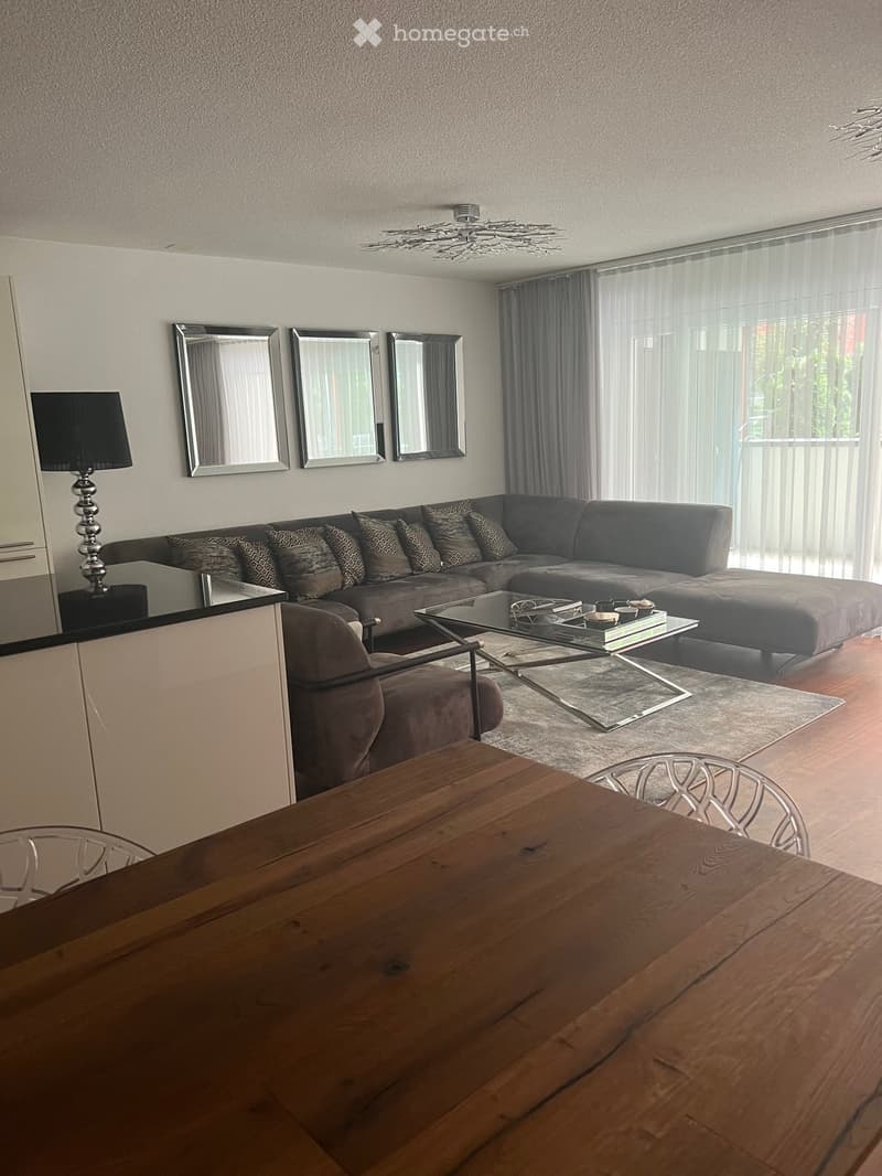 Modern living room with a grey sectional sofa, glass coffee table, wooden dining table, and decorative mirrors on the wall.