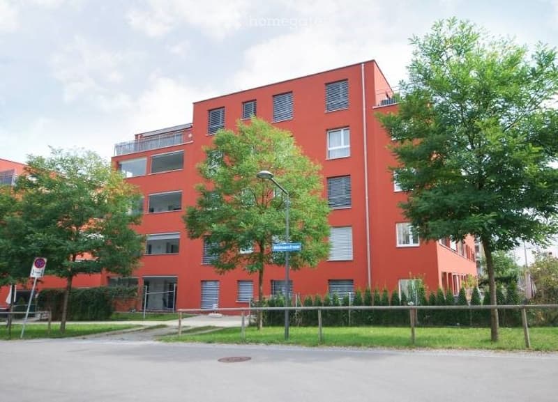 Modern red apartment building with multiple floors, large windows, and balconies. Surrounded by greenery and a paved area.