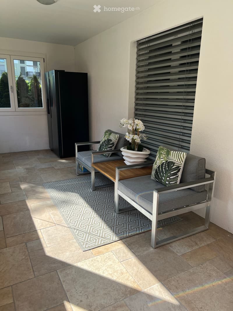 A modern, furnished balcony with a grey sofa, wooden table, decorative rug, and a flower pot with white orchids.