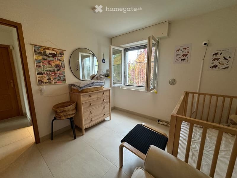A well-lit room with a wooden dresser, a mirror, a small stool, a cradle, and a chair. The room features a large window with blinds and a door leading to another area.