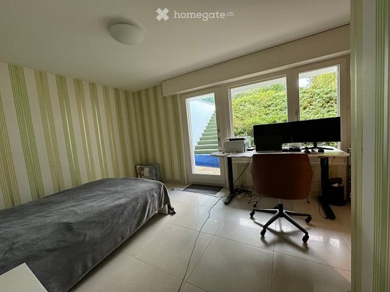 A furnished room with a bed, desk, chair, and a large window with a view of greenery outside. The room features striped wallpaper and tiled flooring.