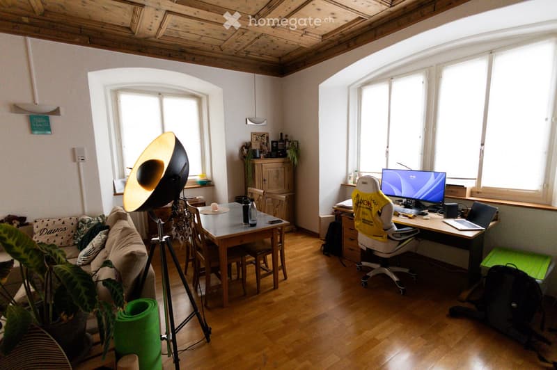 A well-lit room with wooden flooring, featuring a sofa, a dining table with chairs, a desk with a computer and laptop, and a tripod with a large lamp. There is also a cabinet and a plant in the corner.