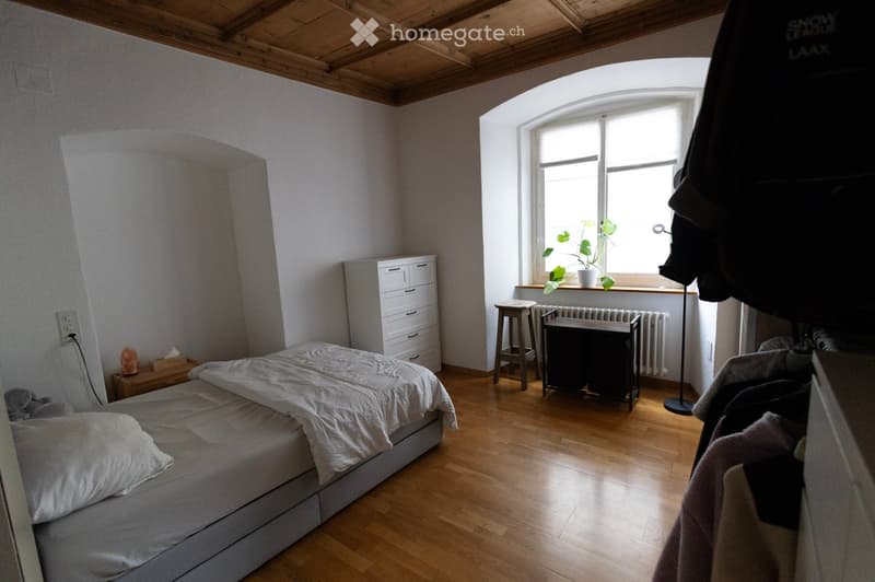Bright bedroom with wooden floor, large window, bed with white bedding, nightstand, drawer unit, small stool, radiator, potted plant on windowsill, and floor lamp.