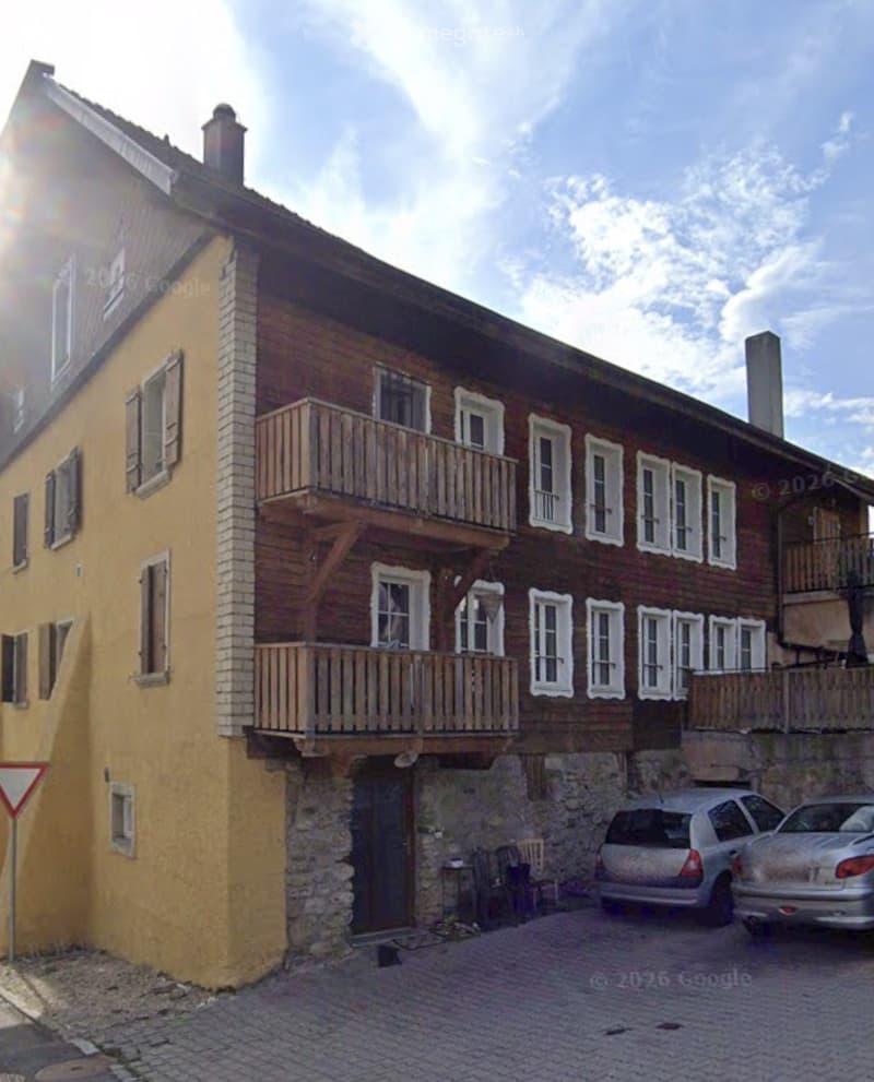 A two-story building with a wooden balcony, stone base, and multiple windows. Two cars are parked in front.