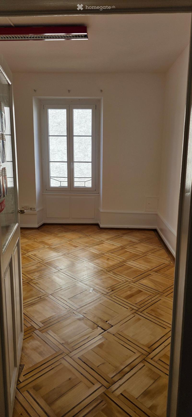 Empty room with wooden herringbone flooring, white walls, and a large window with double doors. No furniture or equipment present.