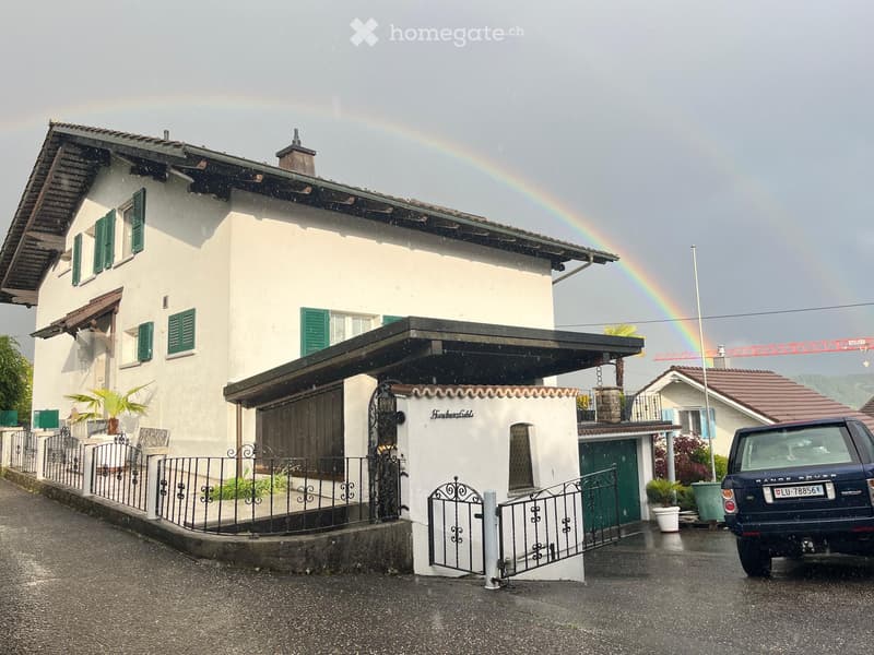 Two-story white house with green shutters, a covered entrance, and a decorative metal gate. A rainbow is visible in the background, and a blue SUV is parked nearby.