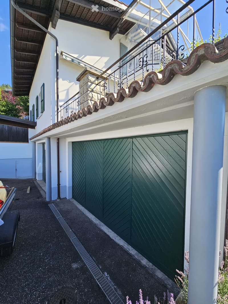 A two-story white building with a green garage door, decorative wrought iron balcony railing, and a stone staircase leading to the upper level.