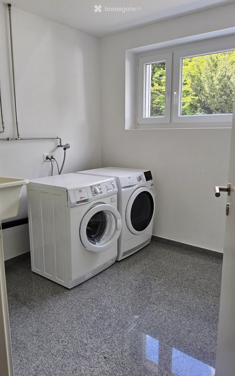 A small, well-lit room with a washing machine and a dishwasher, featuring a grey speckled floor and white walls.