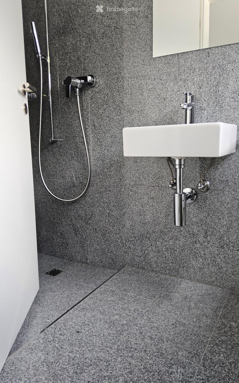 Modern bathroom with a sleek, minimalist design. Features a white sink with a chrome faucet, a showerhead with a handheld sprayer, and grey granite tiles on both walls and floor.