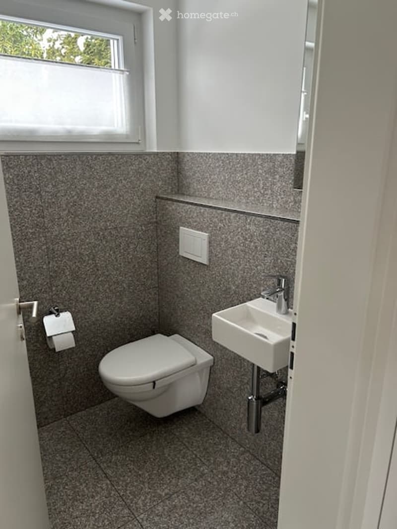 Modern bathroom with a toilet, sink, and window. Grey tiled walls and floor, white fixtures, and a light switch on the wall.