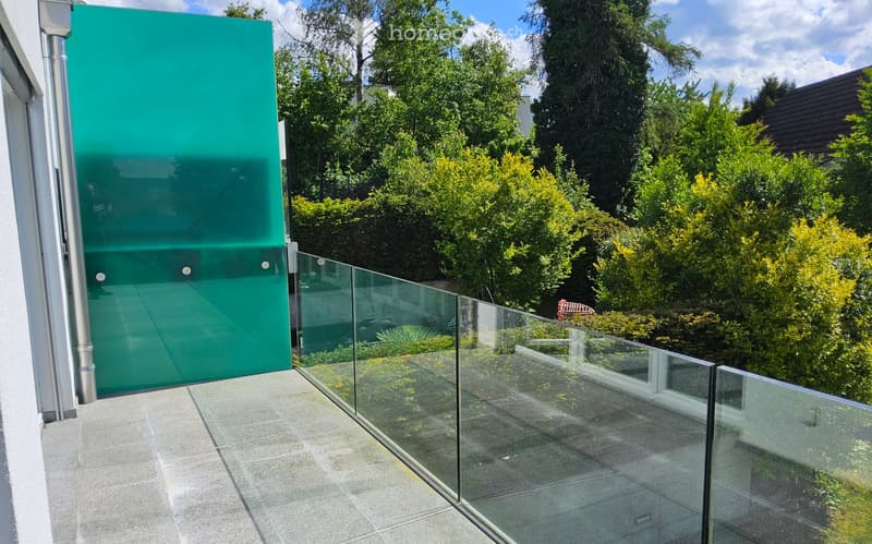 Balcony with glass railing, green screen, and tiled floor, overlooking a lush garden with trees and shrubs.