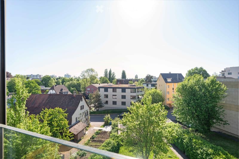 View from a balcony showing a mix of residential buildings, trees, and a small garden area with a pathway.