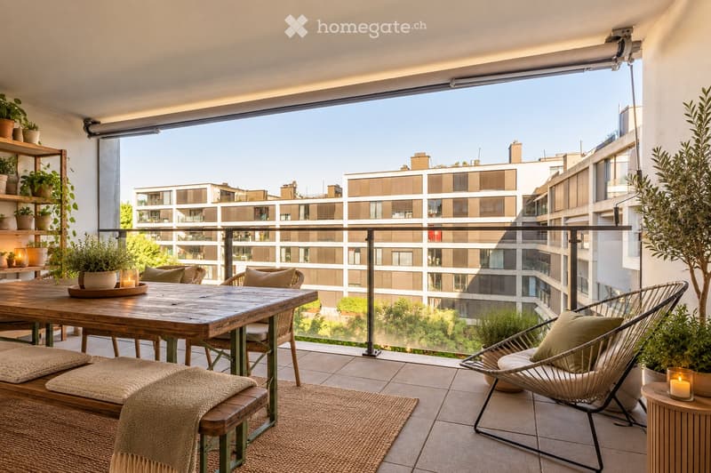 Balcony with wooden table, chairs, bench, cushions, rug, and potted plants, overlooking a modern building complex.